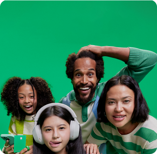 Uma família de quatro pessoas assistindo uma criança jogar vídeo game em um fundo verde, a outra criançå segura um celular e o cartão PicPay para menores.