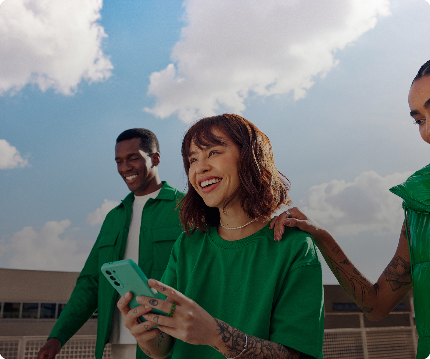 Três pessoas jovens caminham ao ar livre em um dia ensolarado; a pessoa ao centro sorri enquanto segura um smartphone, acompanhada por duas outras pessoas que também parecem animadas.