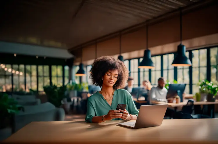 Pessoa em coworking, sentada à mesa com notebook e smartphone, com fundo desfocado.
