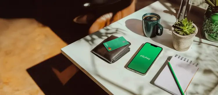 Mesa clara com carteira, cartão PicPay, smartphone com o logo do Picpay na tela, caderno, lápis, caneca e plantas, sob luz natural.