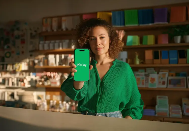 Pessoa em ambiente interno, em frente a prateleiras com diversos produtos, segurando um smartphone à frente do corpo com o logo do PicPay na tela.