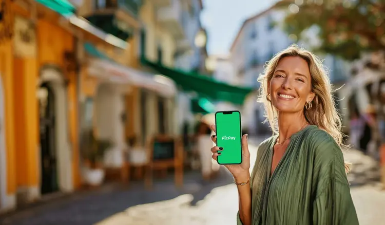 Pessoa em ambiente externo urbano, segurando um smartphone com o logo do PicPay na tela. O fundo mostra uma rua com prédios e fachadas desfocadas