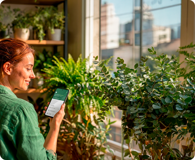 Mulher de blusa verde sorrindo ao olhar o celular em um ambiente cercado por diversas plantas.
