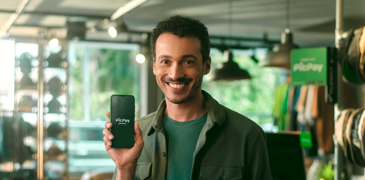 Homem sorrindo vestido com jaqueta verde, segurando um celular com o logo do PicPay Empresas, atrás dele temos um fundo que remete a uma loja de roupas.