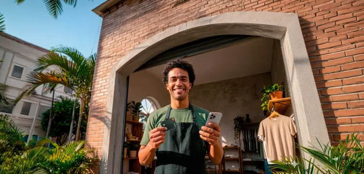 Comerciante sorrindo com cartão e celular na entrada de sua loja.